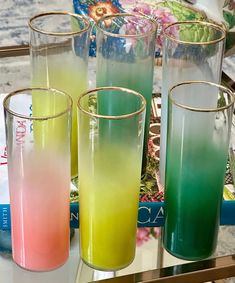 four glasses with different colored liquids on a table next to a book and vases