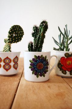 three mugs with different designs on them sitting on a table next to cacti