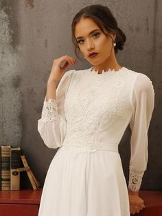 a woman wearing a white dress with long sleeves and lace on the neck, standing in front of a bookcase