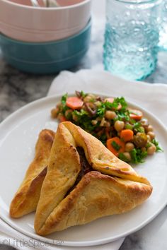 two pastries on a white plate next to a bowl of salad and glass of water