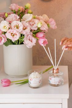 a white vase filled with pink flowers on top of a table next to a candle