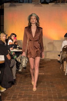 a model walks down the runway in a short brown dress and matching hat with her hair pulled back