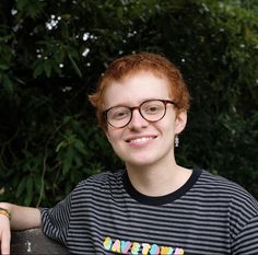 a woman with red hair and glasses sitting on a bench in front of some trees