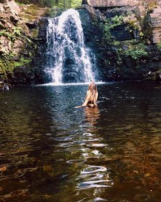 a woman sitting in the water near a waterfall with her back turned to the camera