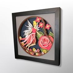 a paper cut fairy surrounded by flowers in a black framed frame on a white wall