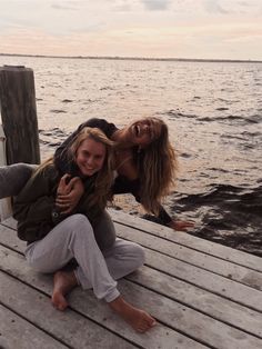 two women are sitting on a dock by the water and smiling at the camera,