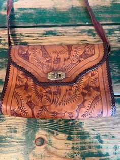 Vintage, tooled leather purse, made in Mexico. This is a small shoulder bag. The purse is tooled with a repeating pattern featuring love bird parrots surrounded by leaves. The purse is primarily a rich caramel brown, with a dark brown braided edge. The purse opens with a top flap. The flap is held down on the front of the bag with a brass twist clasp. Inside, the purse is dyed red. It is separated into two compartments by a zipper pocket in the center. The zipper is stuck where you see it in the Vintage Embossed Satchel For Everyday Use, Vintage Embossed Shoulder Bag For Daily Use, Vintage Brown Hand Tooled Satchel For Daily Use, Vintage Leather Shoulder Bag With Embossed Details, Vintage Leather Embossed Shoulder Bag, Vintage Embossed Leather Shoulder Bag, Vintage Brown Embossed Shoulder Bag, Vintage Brown Embossed Satchel, Brown Embossed Shoulder Bag For Everyday