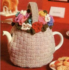 a crocheted tea pot cover with flowers on the top and bottom, sitting next to some cookies