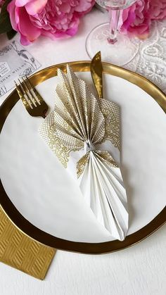 a white plate with gold trim and napkins on it, next to pink flowers