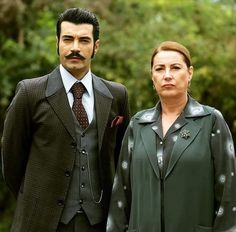 a man and woman standing next to each other in front of some green bushes with trees behind them