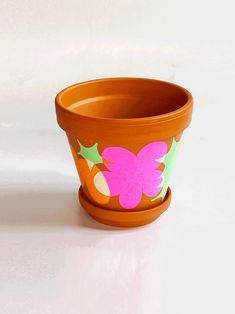 an orange flower pot with pink flowers and green leaves on the rim, sitting on a white surface