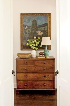 an old dresser with flowers on top and a painting hanging above it in a hallway