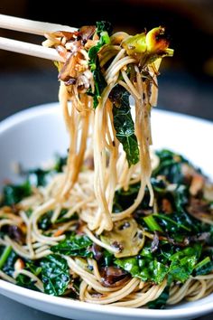 chopsticks holding noodles with greens and mushrooms in a white bowl on a table