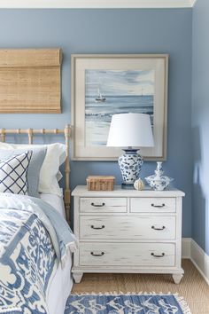 a blue and white bedroom with a painting on the wall next to a dresser, lamp and bed