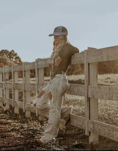 a woman leaning against a fence with her leg up on the rail while wearing jeans and a baseball cap