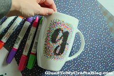 a person holding a coffee mug with sprinkles on it and markers in front of them