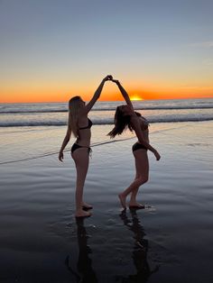 two women in bikinis standing on the beach at sunset with their hands up to each other