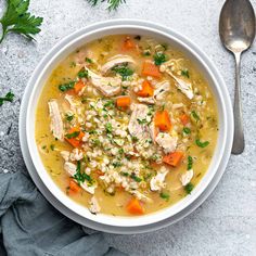 a white bowl filled with chicken and barley soup next to a spoon on a gray surface