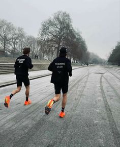 two men in black jackets are running down the street on snow - covered ground with trees and steps behind them