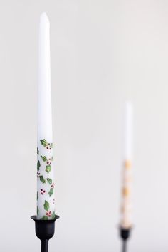 two white candles with holly designs on them sitting side by side in front of each other