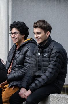 two young men sitting next to each other on a cement wall and looking at the camera