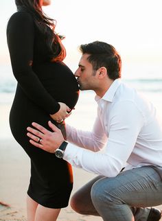 a man kneeling down to kiss a pregnant woman's belly on the beach at sunset