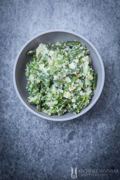 a white bowl filled with green vegetables on top of a gray countertop next to a spoon