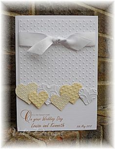 a wedding card with hearts on it and a white ribbon around the edge, in front of a brick wall