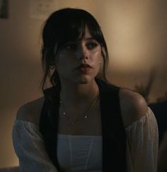 a woman in white shirt and black vest sitting on couch next to wall with lamp