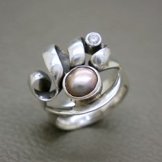 Elevate your jewelry collection with this exquisite vintage sterling silver ring, featuring a stunning natural tinted pearl at its center. Crafted in the 1980s, this ring showcases a unique modernist design that is both bold and elegant, making it a perfect statement piece for any occasion. The sterling silver band is intricately crafted, featuring a unique swirling design that elegantly wraps around a lustrous tinted pearl. A smaller accent diamond cut crystal adds a touch of sparkle, enhancing Unique Silver Pearl Ring For Anniversary, Handmade Vintage Pearl Ring In Sterling Silver, Handmade Vintage Sterling Silver Pearl Ring, Vintage Silver Pearl Ring For Anniversary, Unique Oval Pearl Ring For Anniversary, Vintage Modernist Jewelry, Modernist Jewelry, Modernist Design, Vintage Sterling Silver Rings