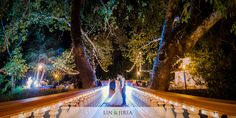 a bride and groom are standing on a bridge at night with lights strung from the trees