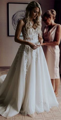 two women standing in front of a window looking at something on the floor while one woman is wearing a wedding dress