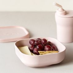 a pink bowl filled with grapes and crackers next to a container full of cheese