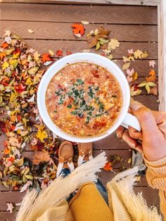 a person holding a bowl of soup in front of autumn leaves on the ground with their hands