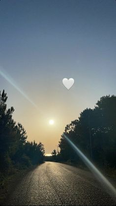 the sun is setting on an empty road with trees in the foreground and a heart - shaped object above it