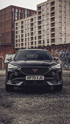 the front end of a black car parked on top of a parking lot next to tall buildings