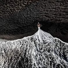 a woman in a white wedding dress standing on top of a wave covered beach with black sand