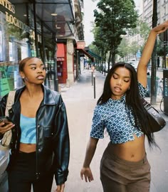 two young women walking down the street in front of a store window, one holding her hand up