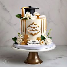 a graduation cake decorated with gold and white icing on a marble pedestal, topped with flowers