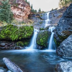 a small waterfall in the middle of some rocks and trees with green moss growing on them