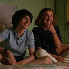 two people sitting on a bed smiling and looking at the camera