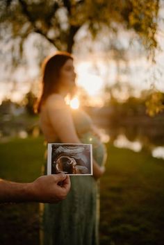 a pregnant woman holding an old photo in front of her belly while the sun is setting