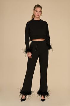 A woman gracefully wears the Rada Feather Trim Pant - Black, featuring stylish ostrich feather trim on the sleeves and pant legs. Posed against a simple beige background, she pairs her ensemble with sleek black heels and sports a chic half-up hairstyle, effortlessly radiating charm. Chic Feather Trim Bottoms For Night Out, Chic Evening Bottoms With Feathers, Chic Feathered Bottoms For Evening, Chic Feather Trim Bottoms, Chic Pants With Feather Trim, Ostrich Feather Trim, Feather Trim, Ostrich Feather, Terry Fabric