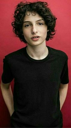 a woman with curly hair standing in front of a red wall wearing a black shirt