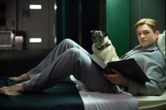 a man sitting on a bed reading a book with a pug in his lap