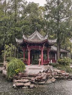 a woman standing in front of a red gazebo on top of a lake surrounded by trees