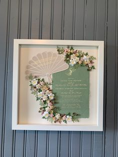 a white frame with flowers and a green paper fan hanging on the wall next to it