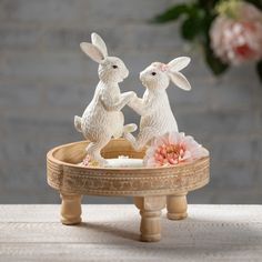 two white rabbits sitting on top of a wooden tray with flowers in front of it