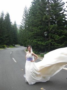 a woman in a wedding dress standing on the side of a road next to trees