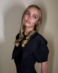 a woman with long blonde hair wearing a black dress and posing for the camera in front of a white wall